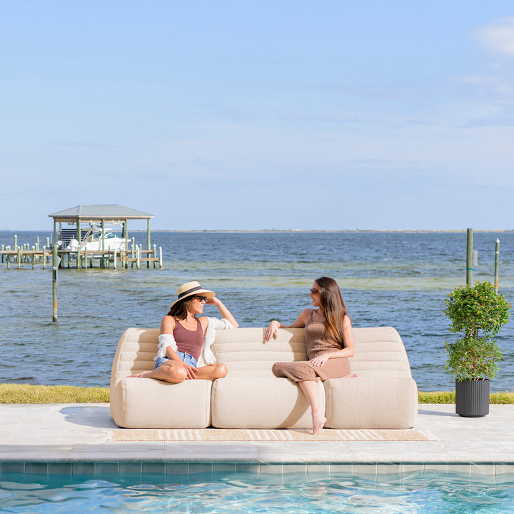Two women sitting on a sofa by a pool with a dock and water in the background #color_sunlit-cream-outdoor-performance-linen