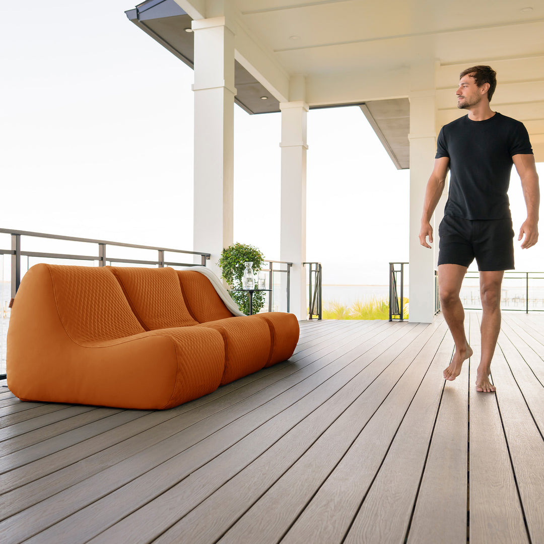 Man walking on a wooden deck with an orange outdoor sofa #color_honey-sand-outdoor-vegan-leather