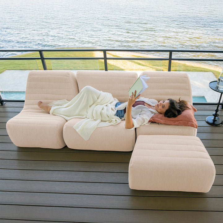 Woman lying on a beige outdoor sofa by a waterfront #color_sunlit-cream-outdoor-performance-linen