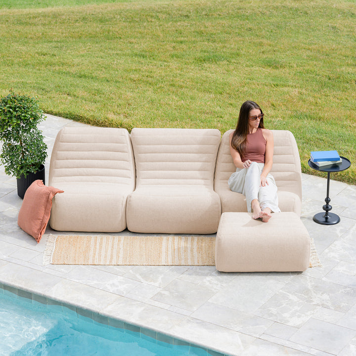 Woman sitting on a beige outdoor sofa by a pool with a grassy area in the background #color_sunlit-cream-outdoor-performance-linen