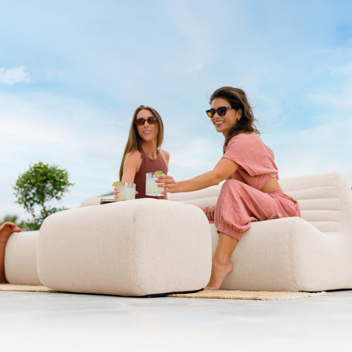 Two women sitting on a beige outdoor sofa with drinks, enjoying a sunny day. #color_sunlit-cream-outdoor-performance-linen