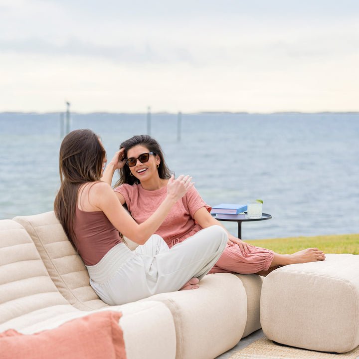 Two women sitting on a beige outdoor sofa by the water, enjoying each other's company. #color_sunlit-cream-outdoor-performance-linen