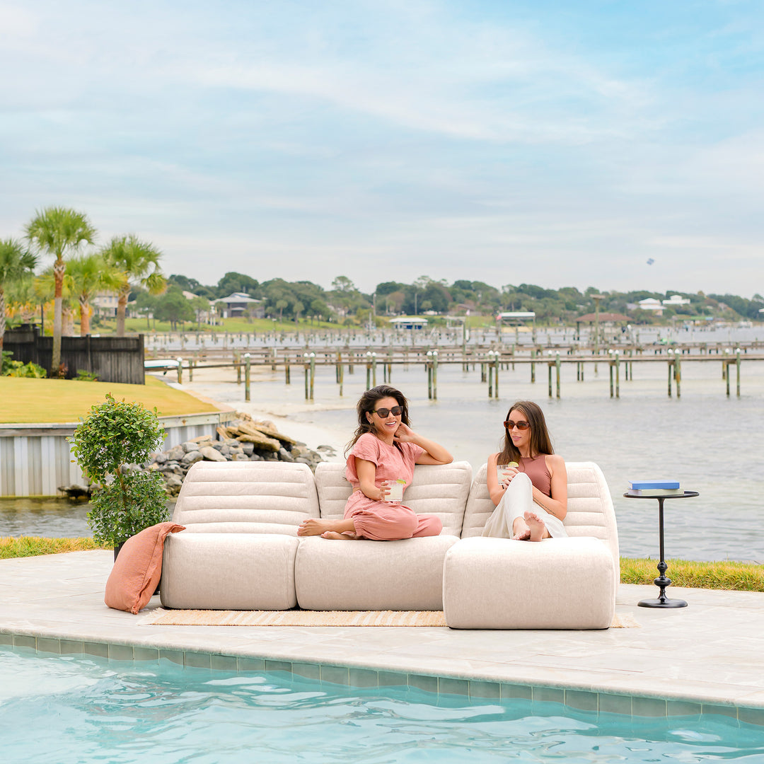 Two women sitting on a beige outdoor sofa by a pool with a waterfront view. #color_sunlit-cream-outdoor-performance-linen