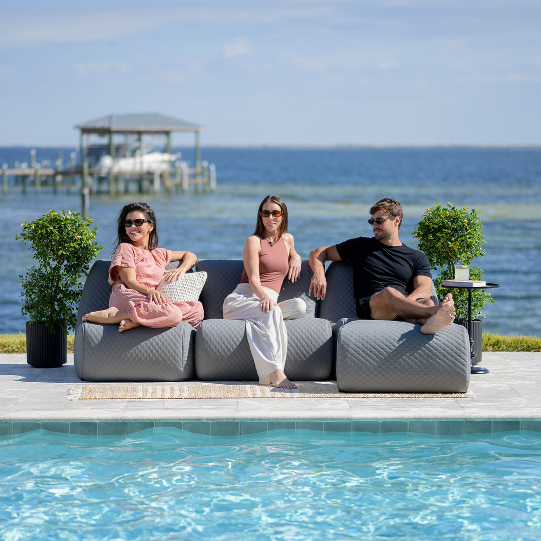 Three people sitting on a modern outdoor sofa by a pool with a scenic background. #color_moonlit-gray-outdoor-vegan-leather
