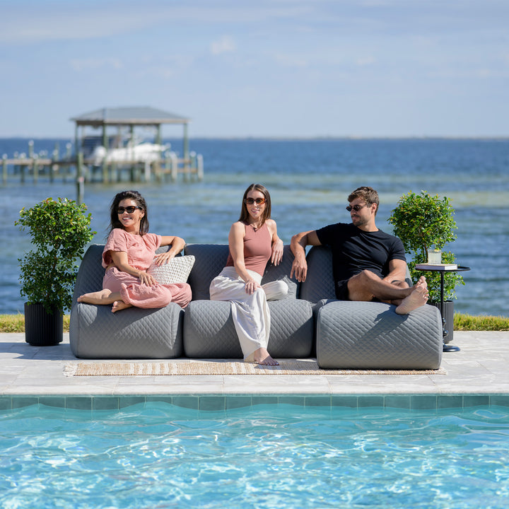 Three people sitting on a modern outdoor sofa by a pool with a scenic background. #color_moonlit-gray-outdoor-vegan-leather
