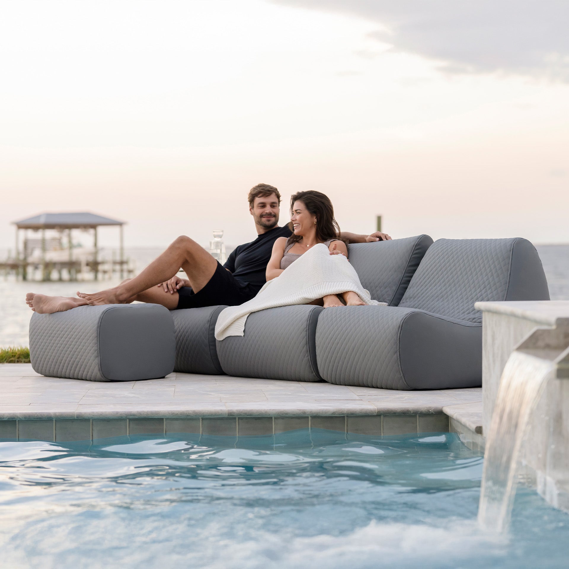 Two people relaxing on a modern outdoor sofa by a poolside. #color_moonlit-gray-cool-touch-silicone-stitched-leather