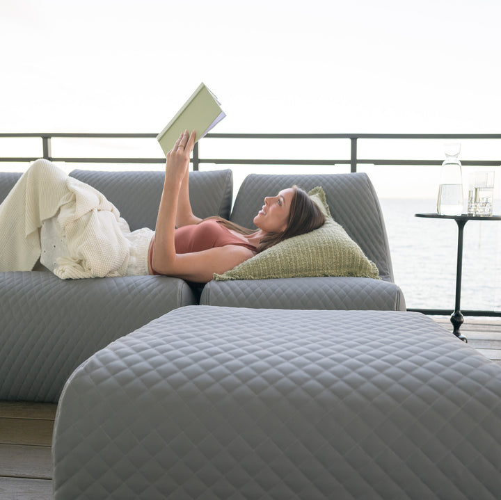 Woman reading a book on a gray sofa with a view of a waterfront. #color_moonlit-gray-outdoor-vegan-leather