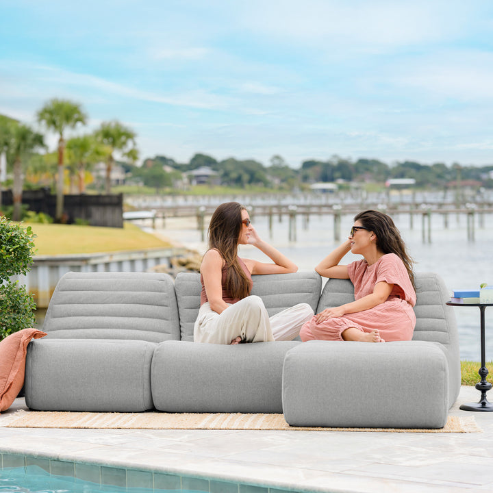 Two women sitting on a gray outdoor sofa by a pool with a waterfront view. #color_port-gray-outdoor-performance-linen
