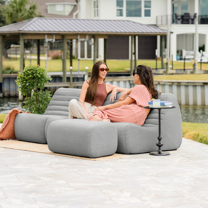 Two women sitting on a gray outdoor sofa by a waterfront with a small table between them. #color_port-gray-outdoor-performance-linen