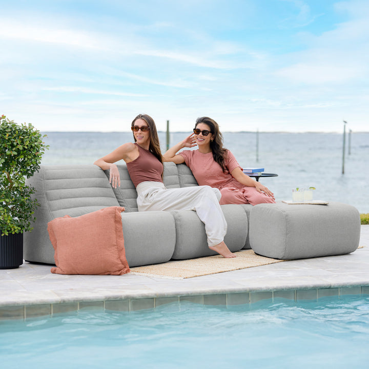 Two women sitting on a gray outdoor sofa by a pool with a scenic background. #color_port-gray-outdoor-performance-linen