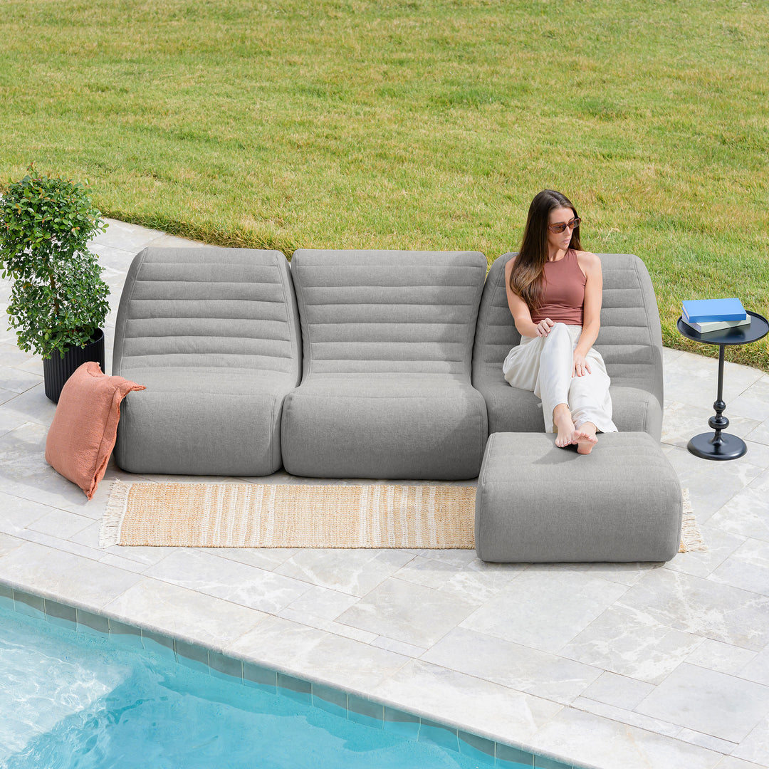 Woman sitting on a gray outdoor sectional sofa by a pool with a grassy area in the background. #color_port-gray-outdoor-performance-linen