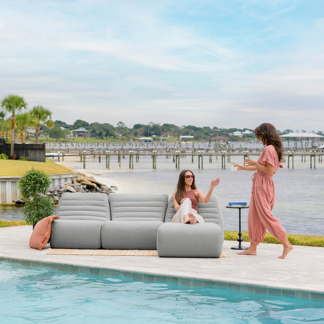 Two women by a pool with a gray sectional sofa and waterfront view. #color_port-gray-outdoor-performance-linen