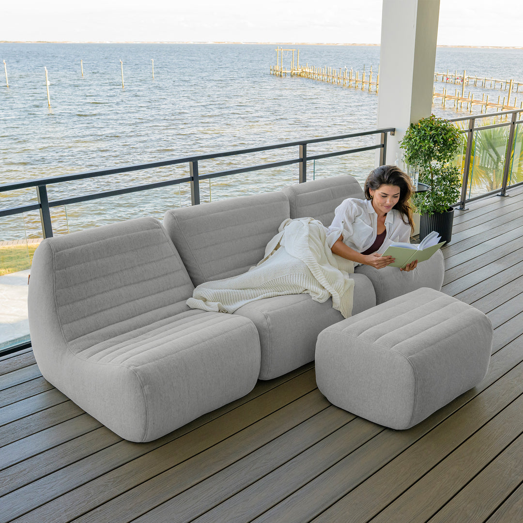 Woman sitting on a modern outdoor sofa with a view of water and a dock. #color_port-gray-outdoor-performance-linen
