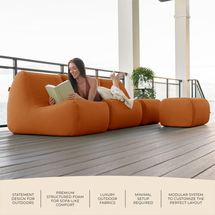 Woman relaxing on an orange outdoor sofa reading a book on a deck. #color_honey-sand-outdoor-vegan-leather