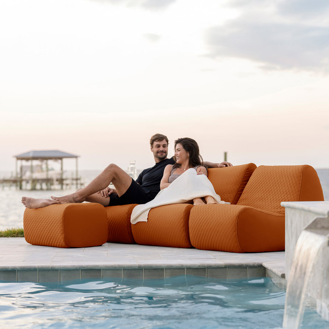 Two people relaxing on an orange outdoor sofa by a pool with a scenic background. #color_honey-sand-outdoor-vegan-leather