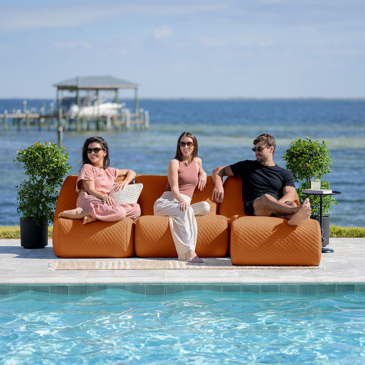 Three people sitting on an orange outdoor sofa by a pool with a scenic background. #color_honey-sand-outdoor-vegan-leather