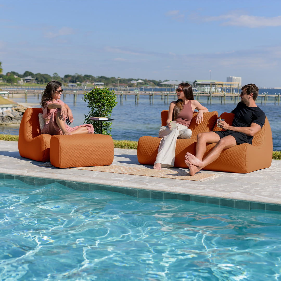 Three people sitting on orange bean bag chairs by a pool with a waterfront view. #color_honey-sand-outdoor-vegan-leather