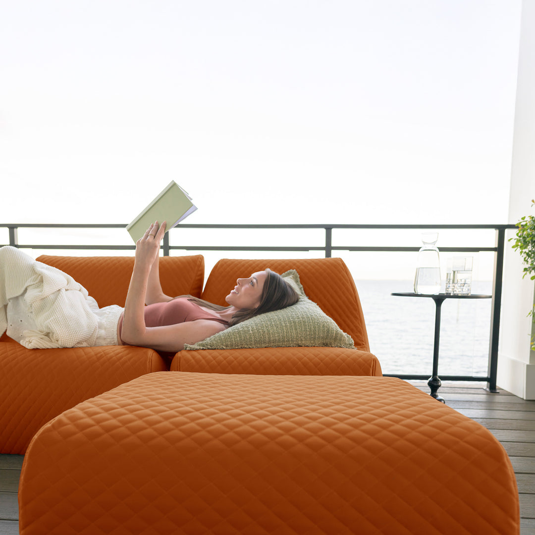 Woman lying on an orange sofa reading a book with a view of the ocean. #color_honey-sand-outdoor-vegan-leather