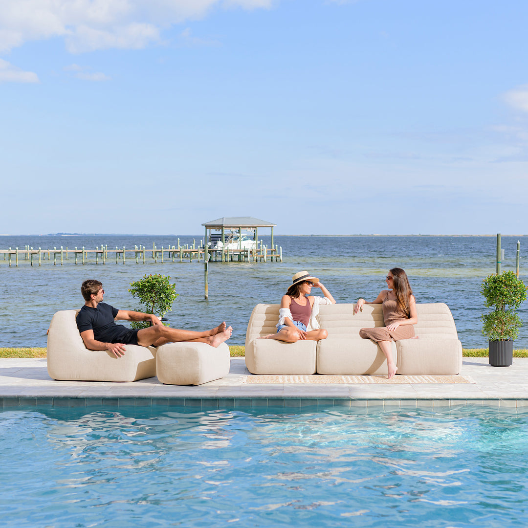 Three people sitting on a poolside sofa with a scenic view of water and sky. #color_sunlit-cream-outdoor-performance-linen