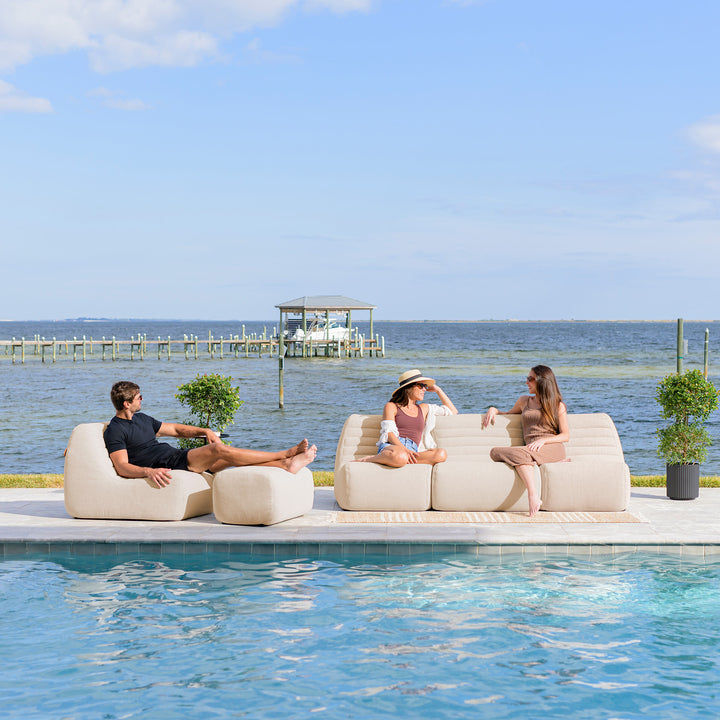 Three people sitting on a poolside sofa with a scenic view of water and sky. #color_sunlit-cream-outdoor-performance-linen