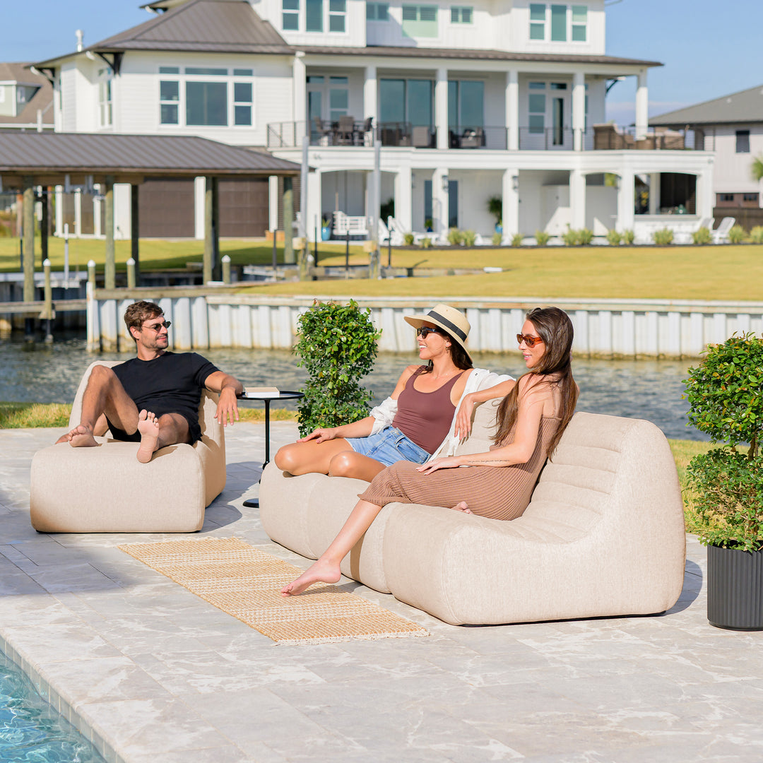 Three people sitting on lounge chairs by a pool with a waterfront house in the background. #color_sunlit-cream-outdoor-performance-linen