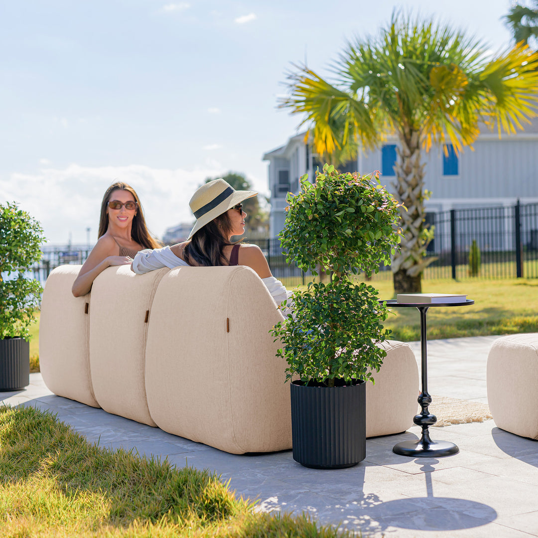 Two women sitting on a beige outdoor sofa with palm trees and a house in the background. #color_sunlit-cream-outdoor-performance-linen