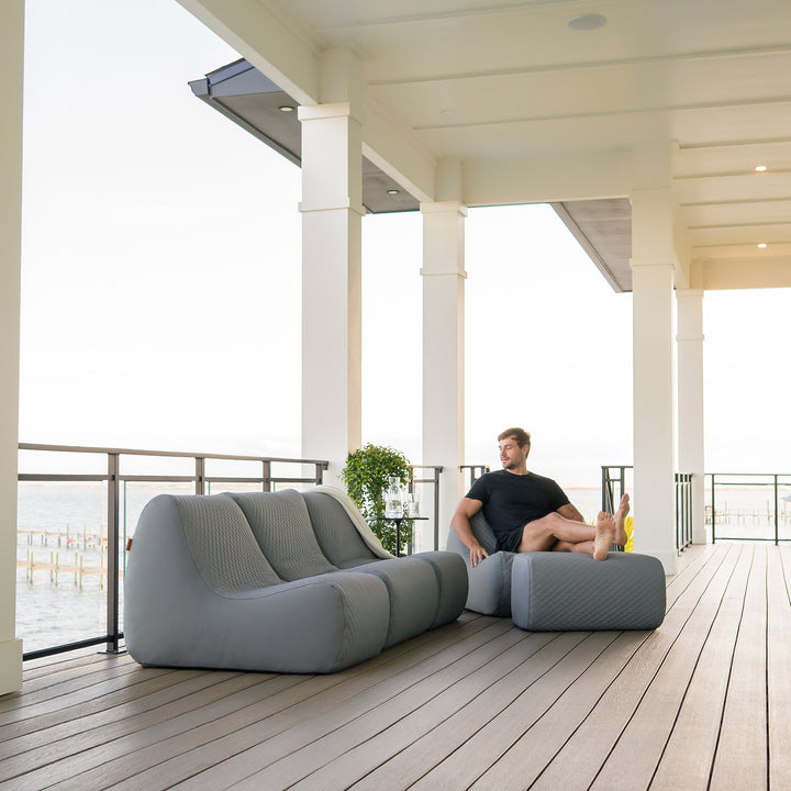 Person sitting on a modern outdoor sofa on a wooden deck with a view of the ocean. #color_moonlit-gray-outdoor-vegan-leather