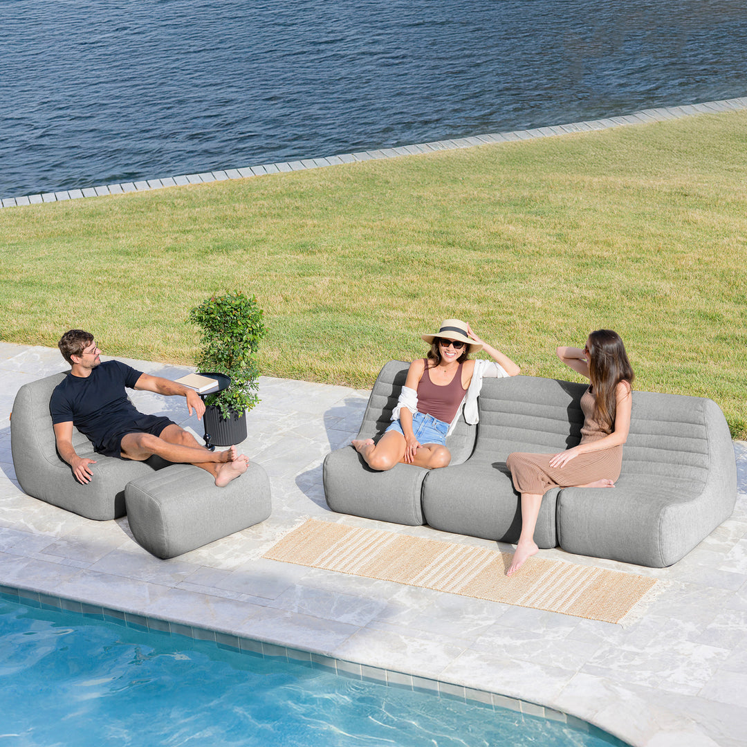 Three people sitting on lounge chairs by a pool with grass and water in the background #color_port-gray-outdoor-performance-linen