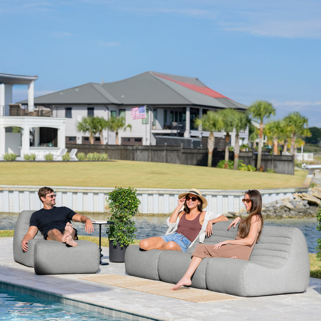 Two women and a man sitting on lounge chairs by a pool with a house and trees in the background. #color_port-gray-outdoor-performance-linen
