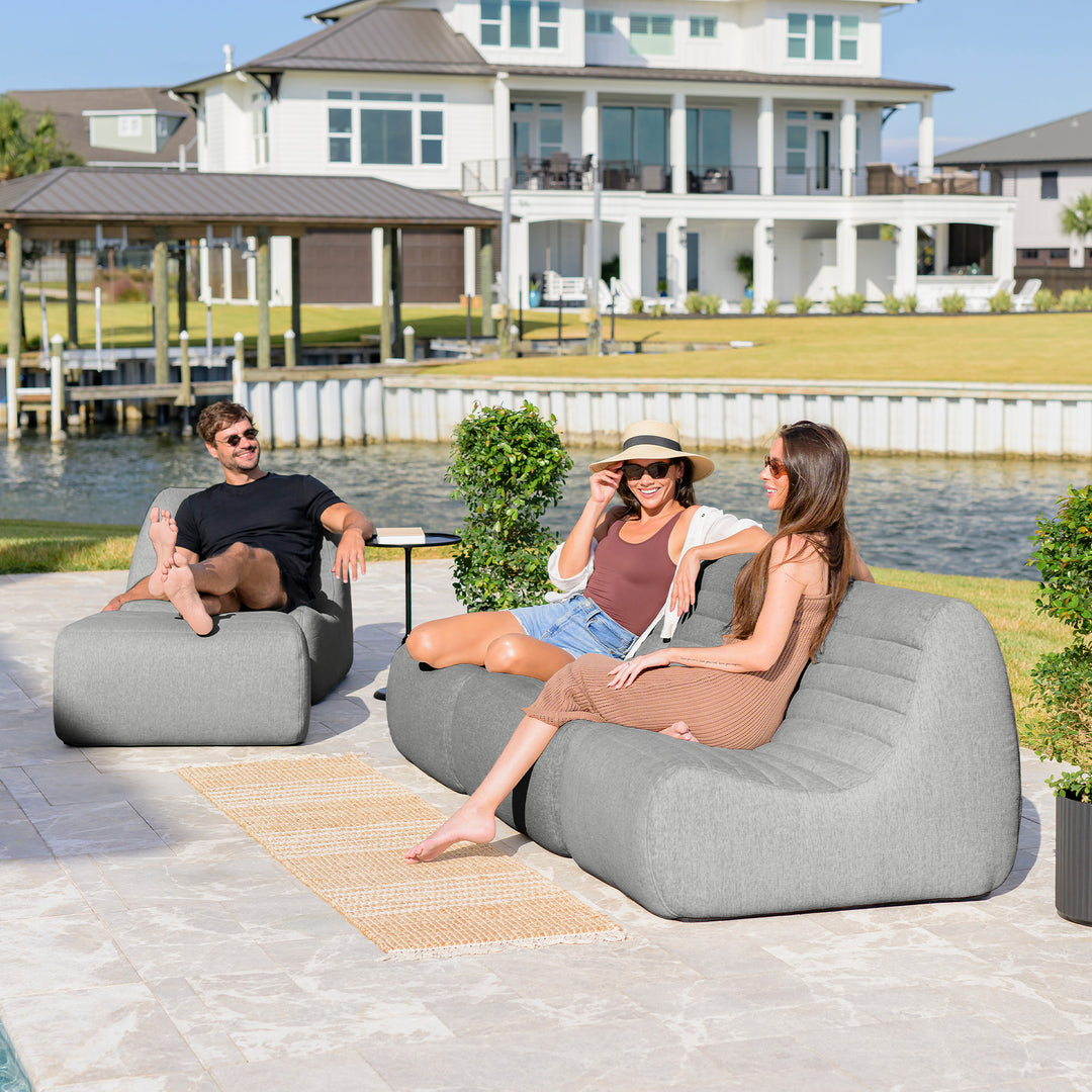 Three people relaxing on lounge chairs by a waterfront with a house in the background. #color_port-gray-outdoor-performance-linen