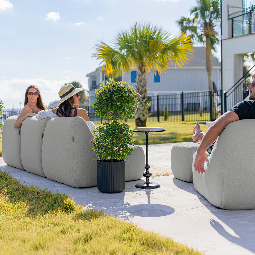Two women and a man sitting on outdoor furniture with palm trees and a building in the background. #color_port-gray-outdoor-performance-linen