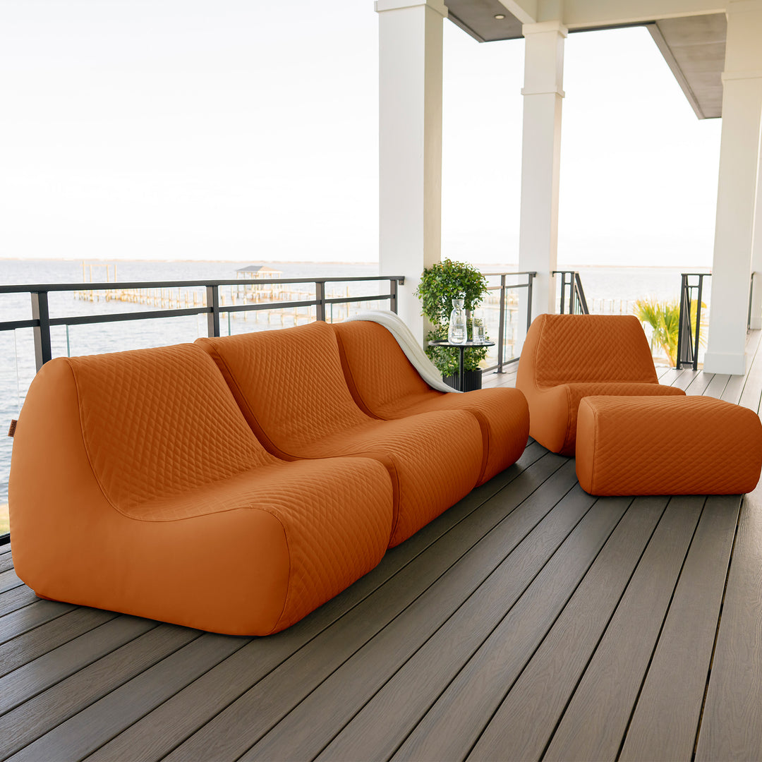 Orange outdoor sofa set on a wooden deck with a white railing and plants in the background. #color_honey-sand-outdoor-vegan-leather