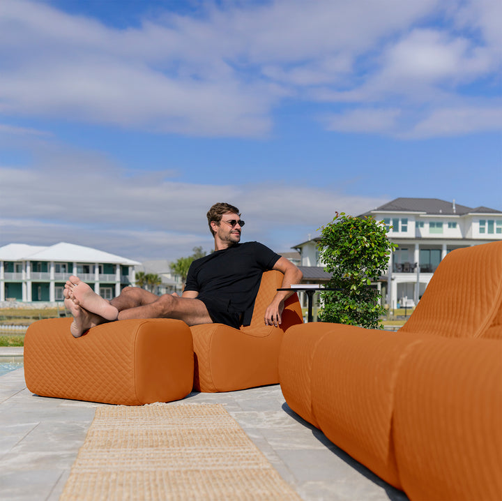 Man sitting on an orange outdoor sofa with a pool and houses in the background #color_honey-sand-outdoor-vegan-leather