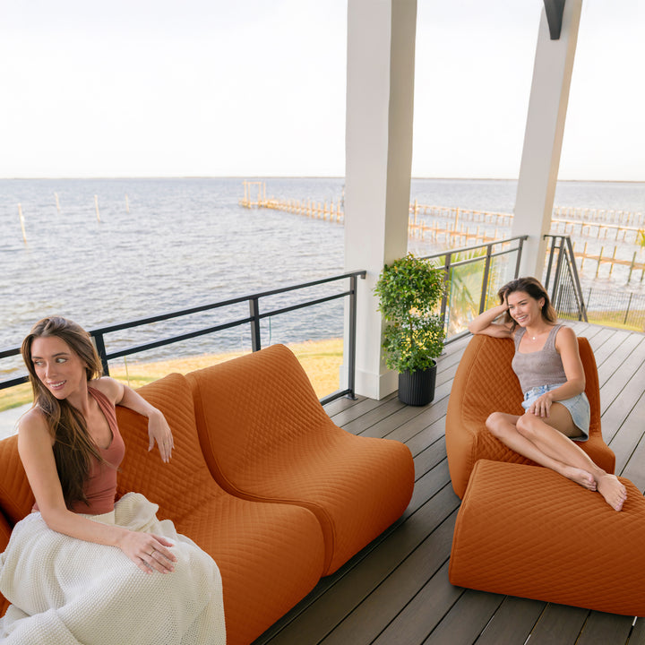 Two women sitting on orange outdoor furniture with a waterfront view. #color_honey-sand-outdoor-vegan-leather