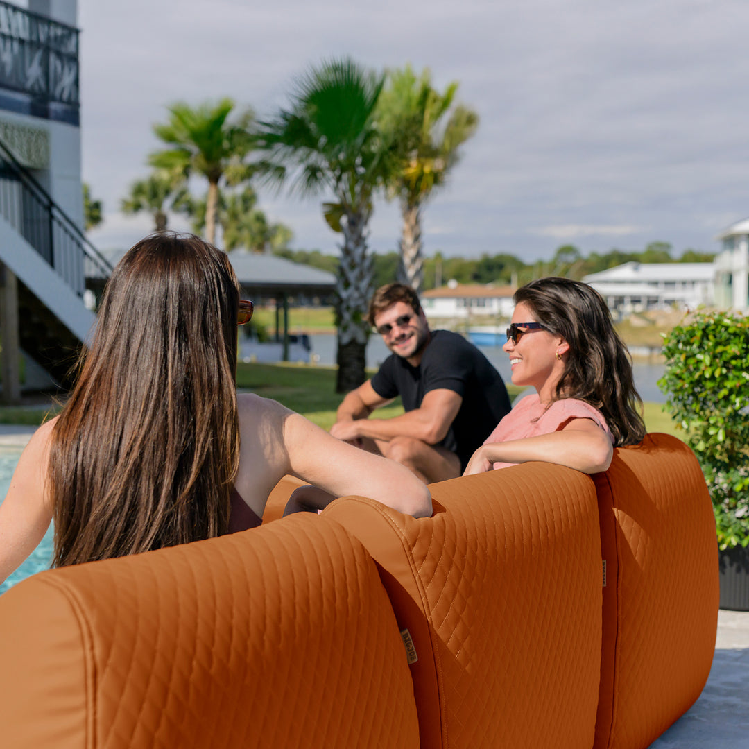 Two women sitting on a brown couch by a pool with palm trees and a building in the background. #color_honey-sand-outdoor-vegan-leather