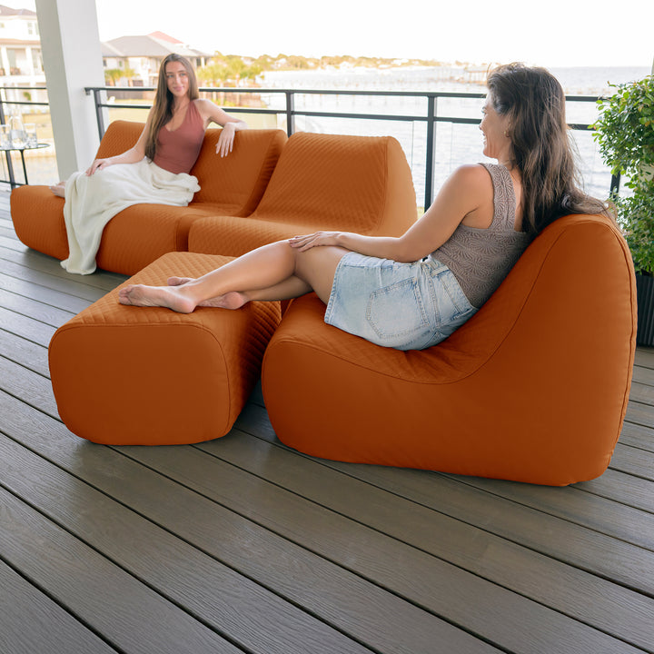 Two women sitting on an orange outdoor sofa on a wooden deck. #color_honey-sand-outdoor-vegan-leather