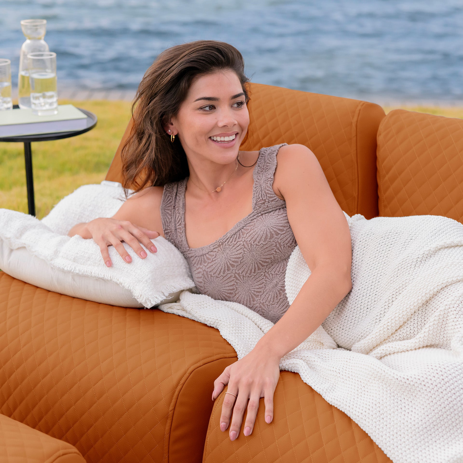 Woman sitting on an orange couch by a body of water, smiling. #color_honey-sand-outdoor-vegan-leather