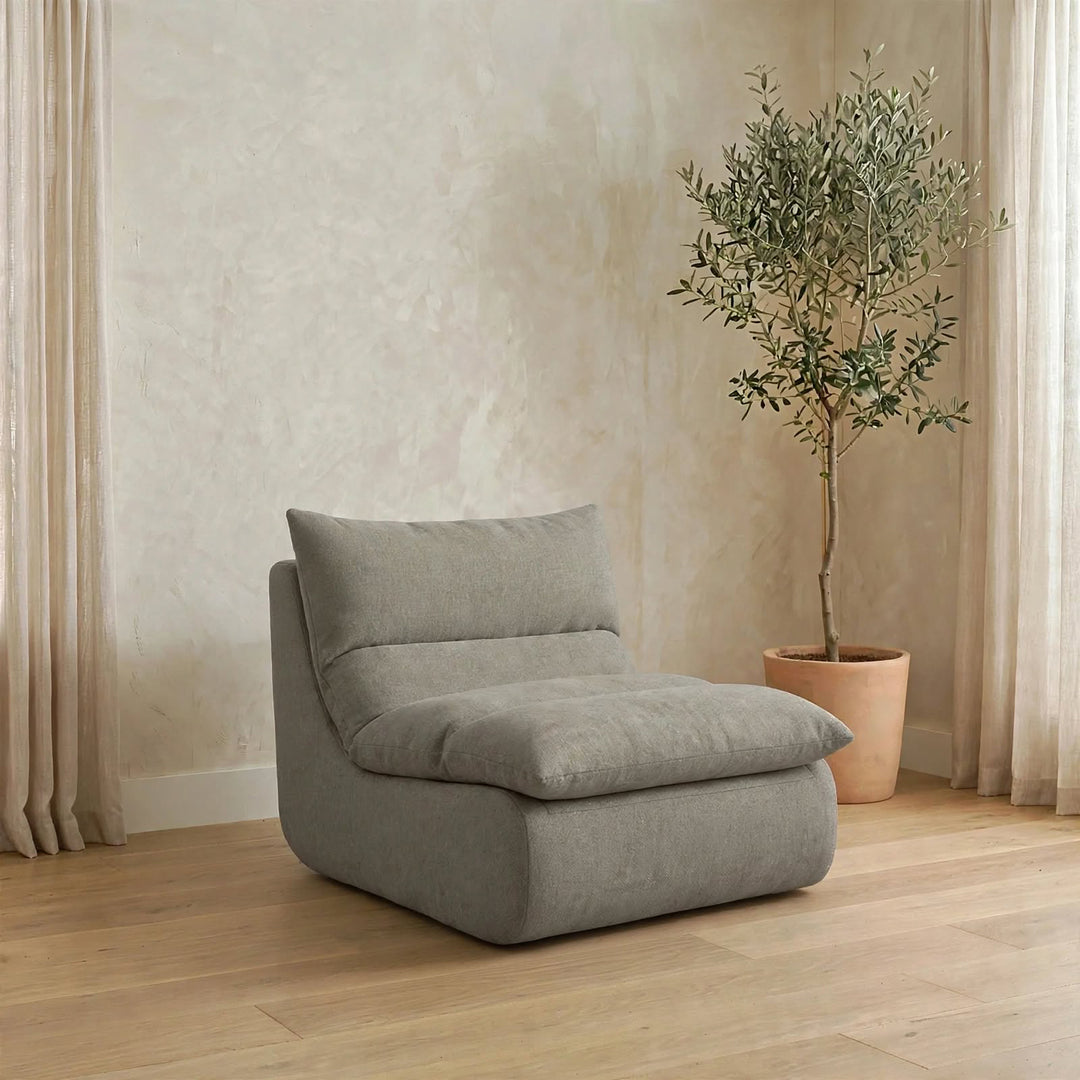 A modern gray upholstered lounge chair with a thick back cushion and padded seat, placed in a softly lit room with wood flooring, beige curtains, and a potted olive tree. #color_misty-gray-terra-knit