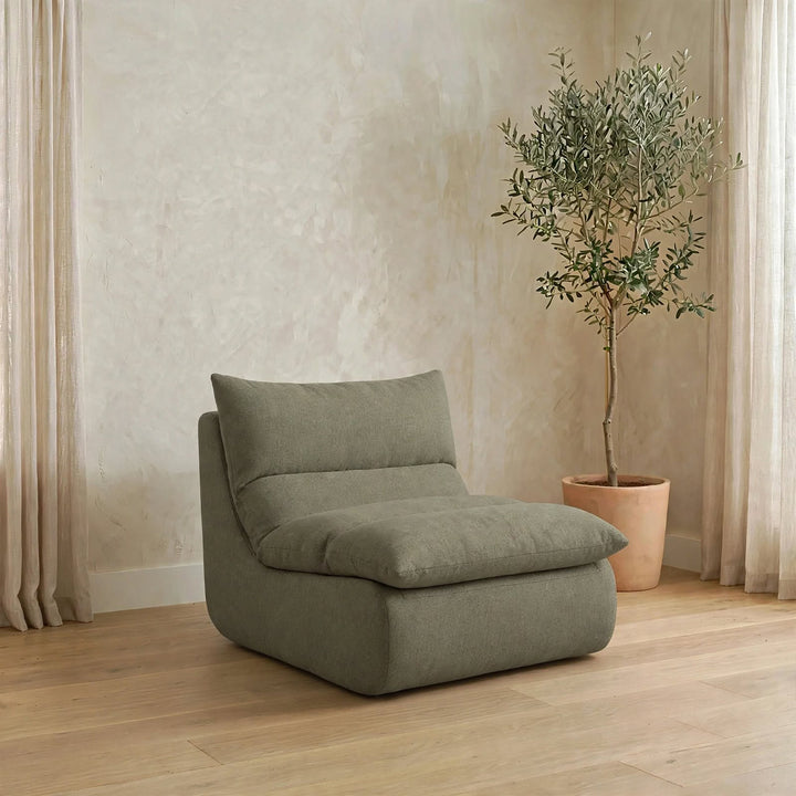 A modern low-profile lounge chair in a muted sage green fabric, styled in a minimalist room with a textured beige wall, light wood flooring, and a potted olive tree.  