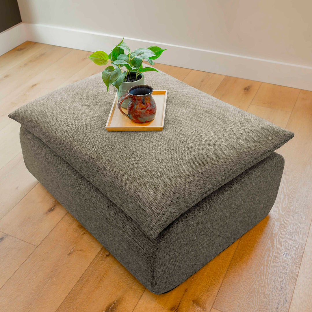 Gray ottoman with a plant, mug, and tray on a wooden floor 