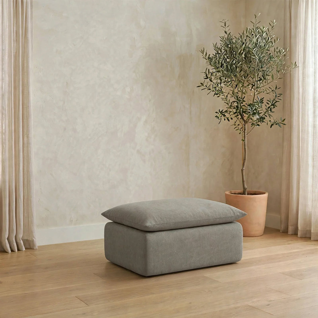 A rectangular ottoman with a matching cushion in a light gray fabric, placed on a light wood floor in a styled room with a potted olive tree and neutral walls. 