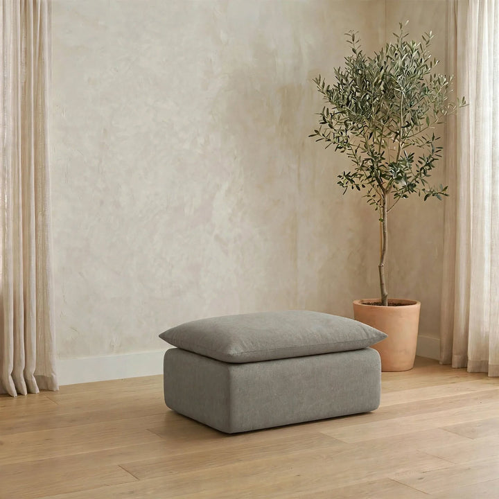 A rectangular ottoman with a matching cushion in a light gray fabric, placed on a light wood floor in a styled room with a potted olive tree and neutral walls. 