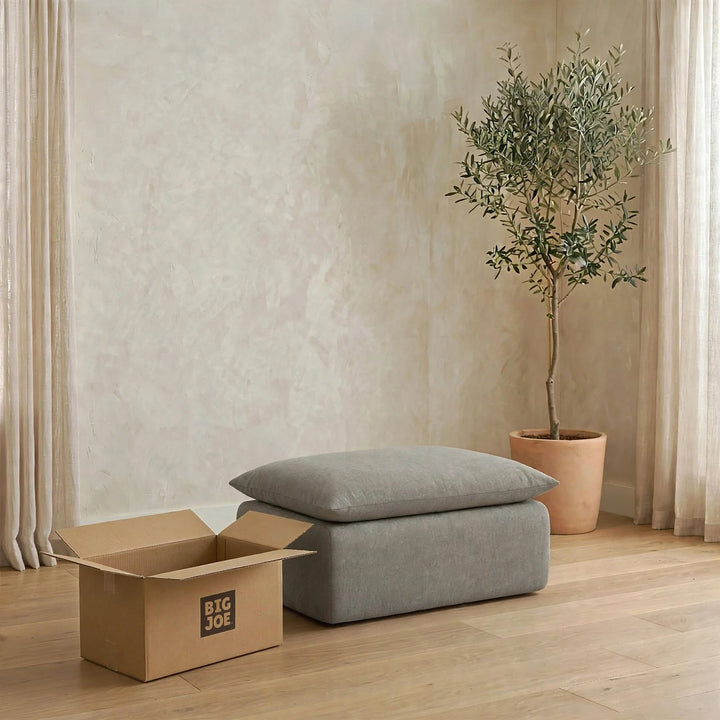 A gray upholstered ottoman or bench with two matching cushions sits on a light wood floor in a styled room. A cardboard box with the visible brand Big Joe is beside it, and a potted olive tree stands against a textured beige wall with sheer curtains. 