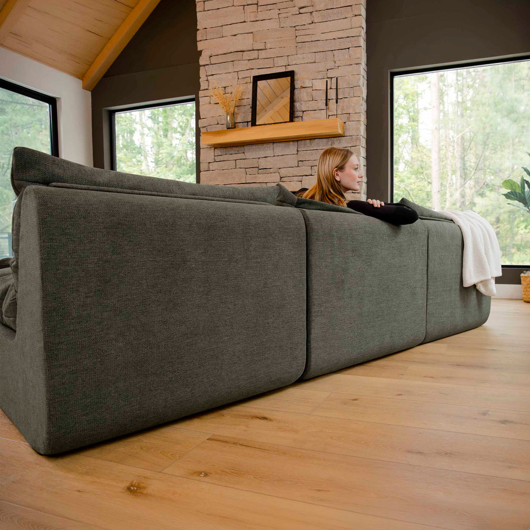 Woman lying on a large gray sofa in a room with a brick wall and large windows. 