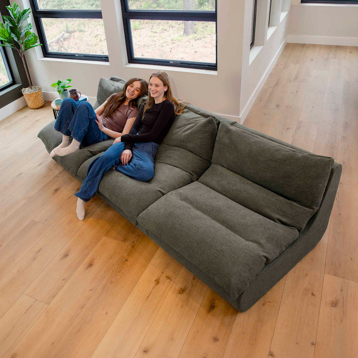 Two women sitting on a large gray sectional sofa in a room with wooden flooring and large windows. 