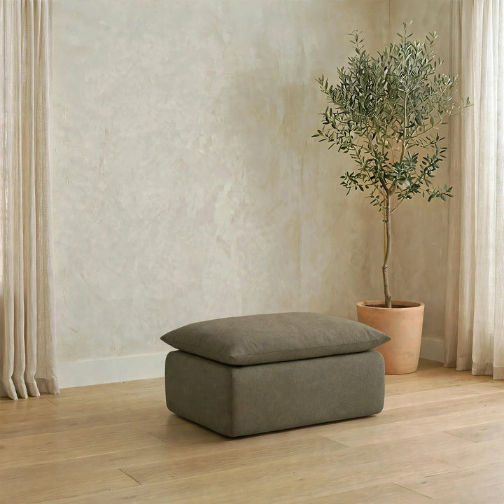 A rectangular ottoman with a cushioned lid in a muted taupe-gray fabric, placed on a light wood floor in a styled room with a potted olive tree and cream curtains.  