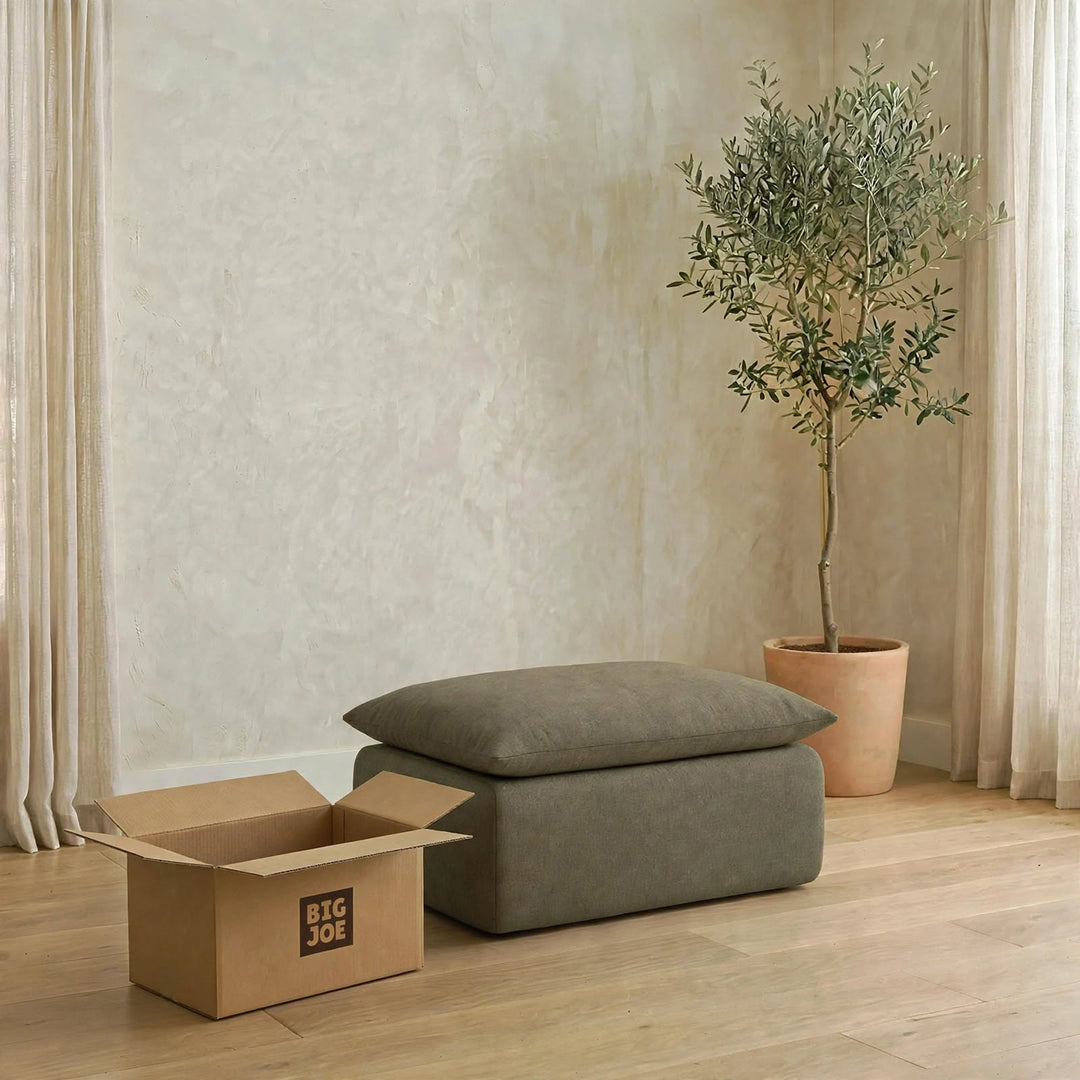 A gray upholstered ottoman with a matching rectangular cushion sits on a light wood floor in a styled room. A large open cardboard box with the visible brand &