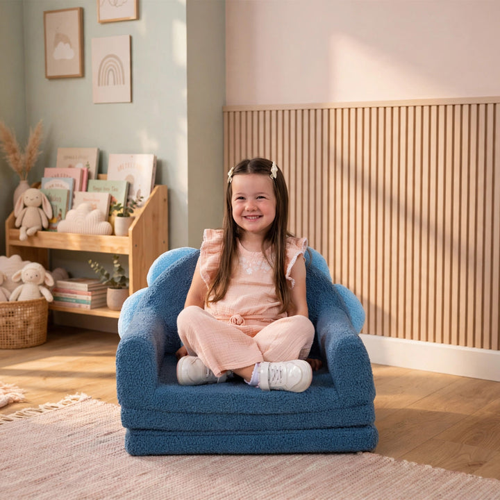 A young girl with long brown hair sits smiling on a blue boucle armchair in a cozy, sunlit children&
