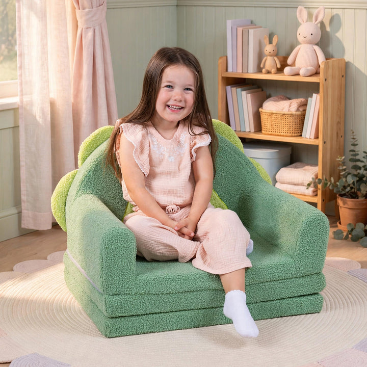 A young girl with long brown hair sits smiling on a small green plush armchair in a cozy nursery. She is wearing a light pink pajama set and white socks. 