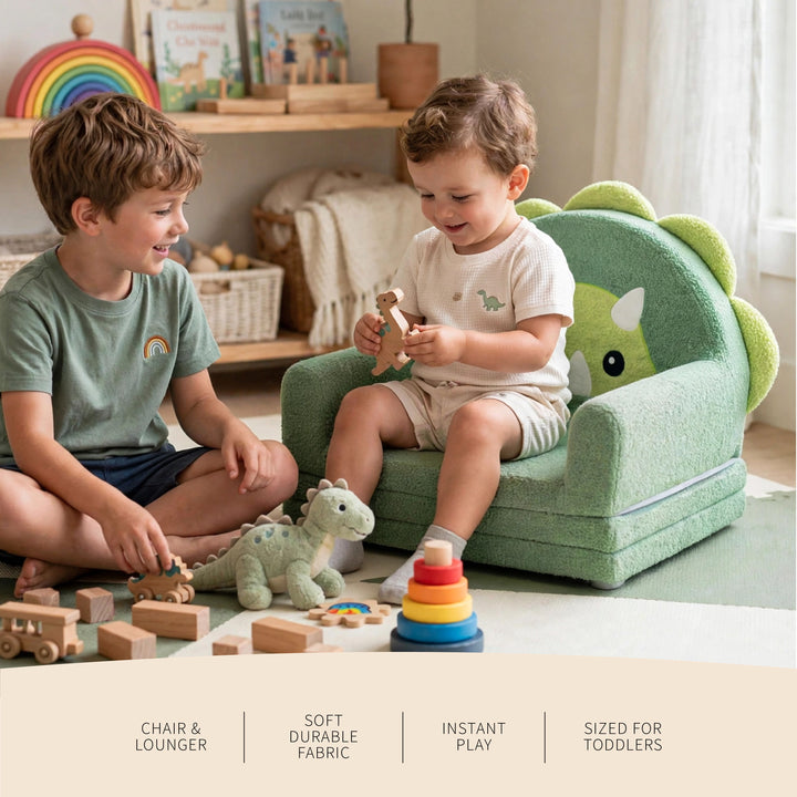 Two toddlers play in a bright nursery or playroom, with one sitting in a green dinosaur-themed chair lounge and the other beside him with toys. The chair has a soft plush fabric, rounded arms, and a dinosaur face on the back. 